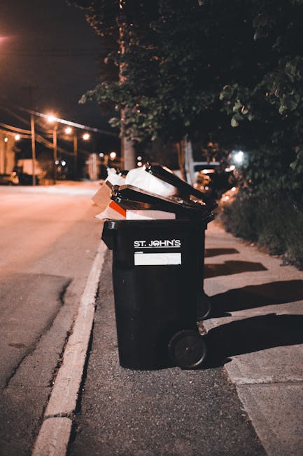 A black plastic rubbish bin with the label 'ST. JOHN'S' positioned upright on a sidewalk at night, filled with a variety of waste including flattened cardboard boxes, paper bags, and other General household rubbish. The bin has a lid partially open, revealing the waste inside, and is situated near a curb on a quiet street. The surrounding environment includes a row of streetlights casting a warm glow, tall trees with dense foliage providing some shelter over the sidewalk, and a background featuring parked cars and residential houses with illuminated windows. The scene captures an urban setting where independent waste disposal, such as rubbish collection by private waste management services, might take place outside traditional council collection hours. The overall atmosphere is calm, and the scene emphasizes the importance of proper waste handling in the context of rubbish removal or alternative disposal options.
