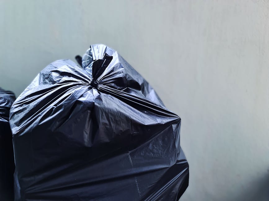 A black plastic rubbish bin with the label 'ST. JOHN'S' positioned upright on a sidewalk at night, filled with a variety of waste including flattened cardboard boxes, paper bags, and other General household rubbish. The bin has a lid partially open, revealing the waste inside, and is situated near a curb on a quiet street. The surrounding environment includes a row of streetlights casting a warm glow, tall trees with dense foliage providing some shelter over the sidewalk, and a background featuring parked cars and residential houses with illuminated windows. The scene captures an urban setting where independent waste disposal, such as rubbish collection by private waste management services, might take place outside traditional council collection hours. The overall atmosphere is calm, and the scene emphasizes the importance of proper waste handling in the context of rubbish removal or alternative disposal options.