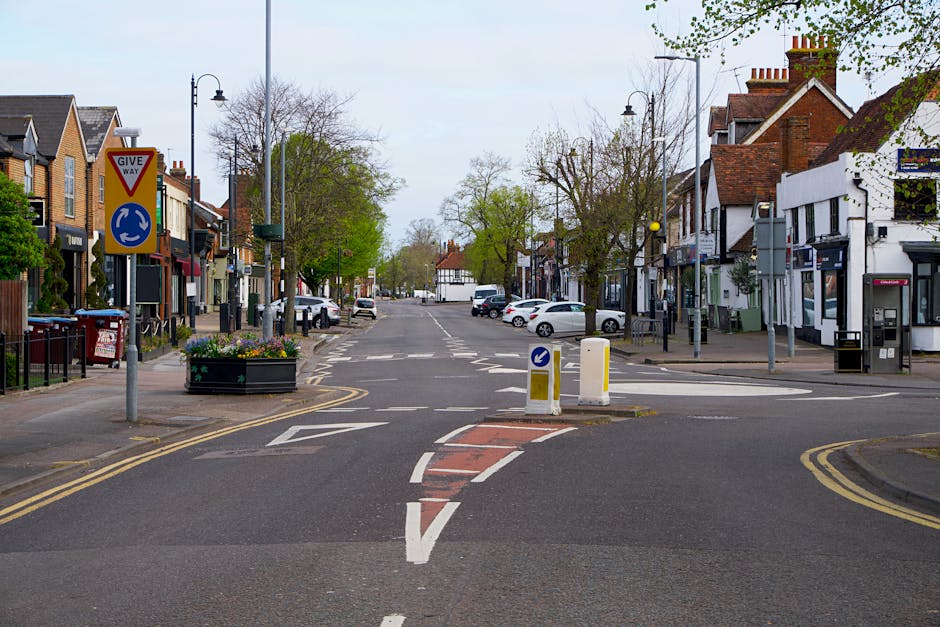 A quiet commercial street scene in a small town, featuring a central paved intersection with white dashed and solid lines, flanked by low-rise buildings including shops and cafes with brick and painted facades. In the foreground, a small island with a black planter containing flowers divides the road, which includes a bollard and directional arrow markings. On the left, a triangle-shaped give way sign is mounted on a pole, alongside street furniture like lampposts, a bus stop, and a red waste bin. The street is lined with parked cars, with some empty spaces further down the street. Tall, mature trees with fresh green leaves are located along the pavement, with additional trees and buildings visible in the background under an overcast sky, creating a calm, well-maintained urban environment suited for local pedestrian and vehicle activity, often associated with community or independent retail areas that may require private rubbish collection services.