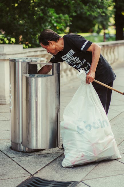 A middle-aged woman with short dark hair, wearing a black T-shirt with geometric white and grey designs, is seen collecting litter and placing it into a large white plastic bag. She is outdoors on a paved sidewalk, leaning slightly forward while holding a wooden litter picker in her right hand. In front of her are three stainless steel and cylindrical litter bins aligned along a low concrete wall, with the bins having open tops for easy waste disposal. The background features lush green trees and bushy foliage, indicating a park or suburban area. The scene is well-lit with natural daylight, highlighting the textures of the stainless steel bins, the woman's clothing, and the paved surface beneath her feet. All elements depict an organized effort of a private individual engaging in rubbish collection or waste management, reflecting an alternative waste handling method aligned with the context of rubbish removal services explained on the webpage.