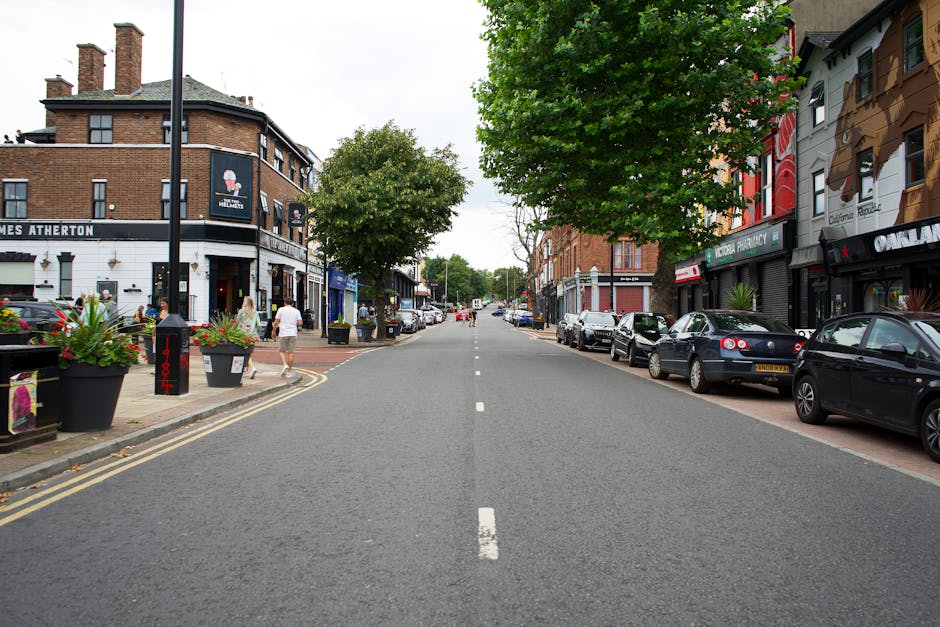A quiet commercial street scene in a small town, featuring a central paved intersection with white dashed and solid lines, flanked by low-rise buildings including shops and cafes with brick and painted facades. In the foreground, a small island with a black planter containing flowers divides the road, which includes a bollard and directional arrow markings. On the left, a triangle-shaped give way sign is mounted on a pole, alongside street furniture like lampposts, a bus stop, and a red waste bin. The street is lined with parked cars, with some empty spaces further down the street. Tall, mature trees with fresh green leaves are located along the pavement, with additional trees and buildings visible in the background under an overcast sky, creating a calm, well-maintained urban environment suited for local pedestrian and vehicle activity, often associated with community or independent retail areas that may require private rubbish collection services.