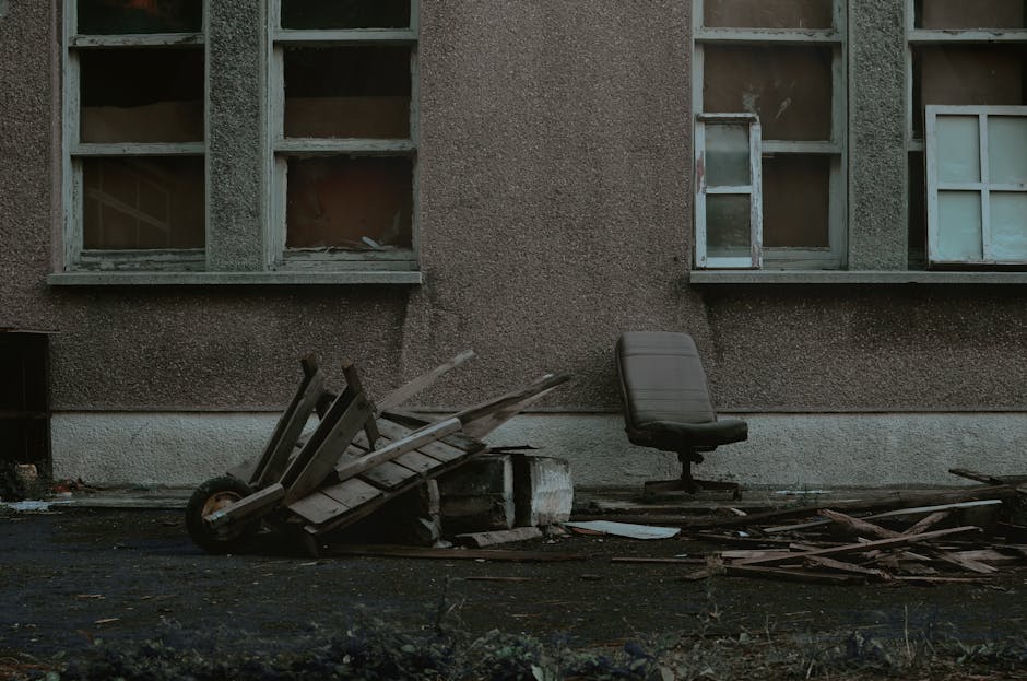 The image depicts an exterior scene adjacent to a weathered, brown-speckled building wall with three large, rectangular windows that have multiple panes, some of which are damaged or missing. In the foreground, there is a collection of discarded debris including a partially tipped-over wooden pallet, which appears aged and weather-beaten with rough, splintered planks. Next to the pallet, a black office chair with a fabric seat and backrest is positioned upright, standing on a wheeled base. To the right, scattered pieces of broken wood and miscellaneous refuse are strewn across the dark pavement, indicating neglect and incomplete clearance. The scene suggests a private or independent rubbish removal attempt, with the debris arranged haphazardly alongside the building, emphasizing the need for professional waste collection services such as those provided by wastecollectioncollierswood.co.uk for efficient on-site clearance of bulky items and household rubbish in the Colliers Wood area.