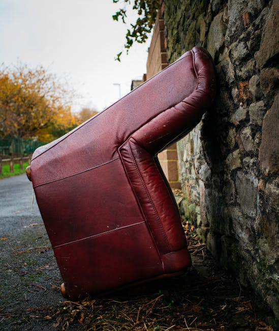 A large, dark red leather sofa with visible creases and a slightly worn surface is positioned upside down and leaning against a rough stone wall on a paved outdoor pathway. The sofa's armrest and backrest are clearly defined, with the textured leather showing some minor scuffs. The setting appears to be a residential area, with trees showing autumn foliage and a partly cloudy sky visible in the background. To the left, a wooden fence lines the pathway, and part of a brick building can be seen behind the sofa. The scene suggests a private waste clearance or alternative rubbish removal, with [COMPANY_NAME] potentially involved in the collection of bulky waste items like furniture pieces being disconnected from their usual interior environment. The ground around the sofa is scattered with small leaves and debris, indicative of outdoor storage or disposal area for unwanted household furniture awaiting collection, consistent with private rubbish removal service practices.