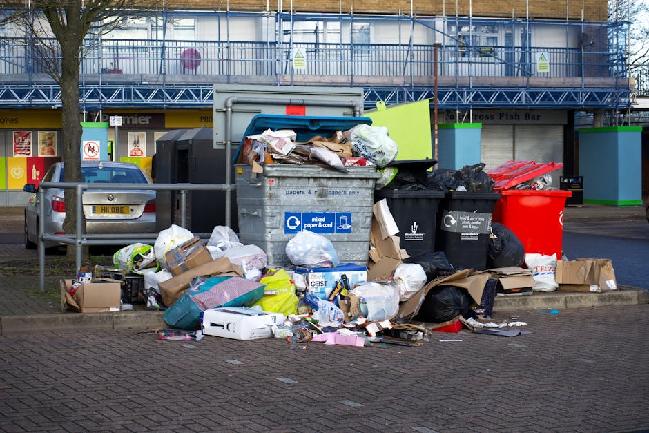 A large collection of mixed waste, primarily composed of paper and cardboard, is piled on an urban sidewalk in front of a building with retail shops and a blue scaffolding-covered upper facade. The waste includes crumpled, flattened cardboard boxes, paper bags, and loose sheets of paper, some torn or partially flattened. Several black and red plastic rubbish bins are positioned behind the pile, with some overflowing and containing black plastic garbage bags. A grey wheelie bin with a blue lid, marked with recycling symbols for paper and card, is prominently visible at the center, containing partially visible waste materials and surrounded by scattered packaging, plastic bags, and smaller cardboard boxes. To the left, a grey car is parked parallel to the waste, with a metal barrier separating the vehicle from the rubbish. The environment appears to be an urban commercial area with paved surfaces, and the scene suggests civilian rubbish accumulation that may require professional waste collection services, such as those offered by wastecollectioncollierswood.co.uk, for effective removal and recycling handling.