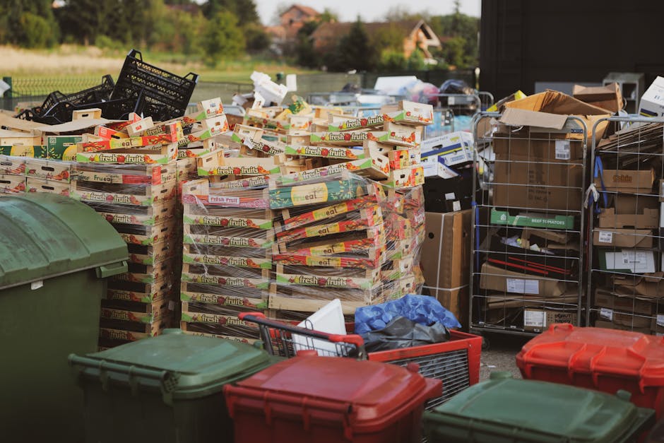 An outdoor scene displaying a large pile of stacked cardboard fruit boxes with colorful printed labels, primarily yellow, red, and green, arranged in a haphazard manner on concrete ground. To the right, there are metal wire cages filled with various cardboard boxes, some partially open, revealing contents like paper and packaging materials. In the foreground, multiple plastic waste bins with green and red lids are visible, some with lids closed and others open, revealing additional rubbish such as plastic bags and miscellaneous debris. The background includes a grassy area with trees and a distant residential building under a partly cloudy sky. The scene suggests an area designated for waste collection or recycling, with an emphasis on cardboard and packaging disposal that can be associated with private rubbish removal services, like those provided by companies such as wastecollectioncollierswood.co.uk.