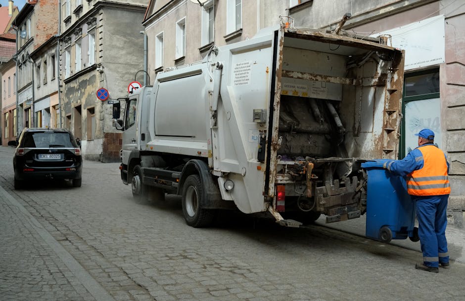 A large collection of mixed waste, primarily composed of paper and cardboard, is piled on an urban sidewalk in front of a building with retail shops and a blue scaffolding-covered upper facade. The waste includes crumpled, flattened cardboard boxes, paper bags, and loose sheets of paper, some torn or partially flattened. Several black and red plastic rubbish bins are positioned behind the pile, with some overflowing and containing black plastic garbage bags. A grey wheelie bin with a blue lid, marked with recycling symbols for paper and card, is prominently visible at the center, containing partially visible waste materials and surrounded by scattered packaging, plastic bags, and smaller cardboard boxes. To the left, a grey car is parked parallel to the waste, with a metal barrier separating the vehicle from the rubbish. The environment appears to be an urban commercial area with paved surfaces, and the scene suggests civilian rubbish accumulation that may require professional waste collection services, such as those offered by wastecollectioncollierswood.co.uk, for effective removal and recycling handling.